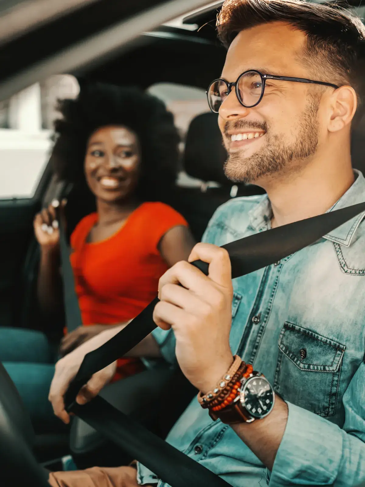 Person fastening a seatbelt in the driver's seat of a car while another passenger sits in the front passenger seat.