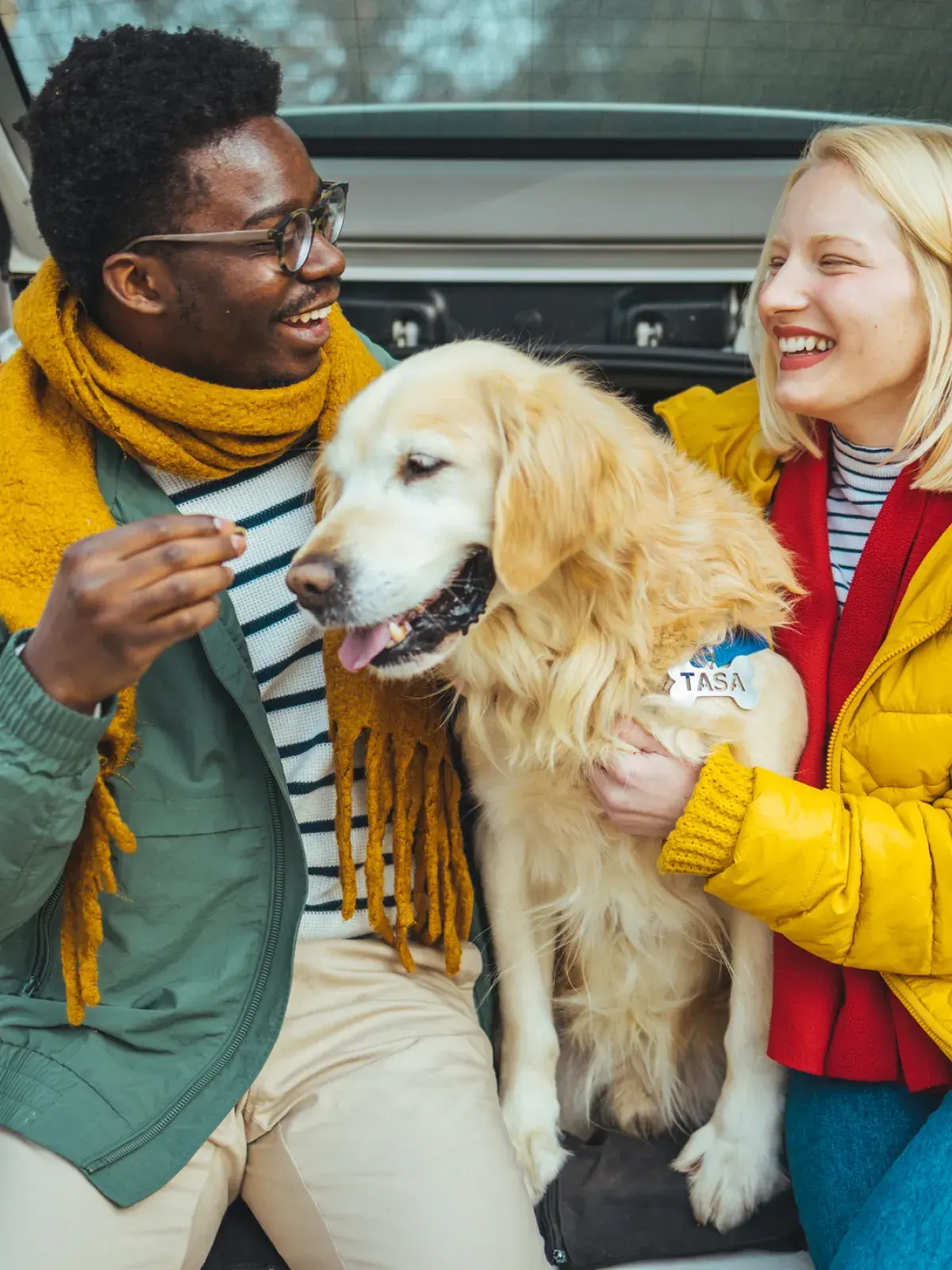 Two people in colorful jackets sit in the open trunk of a parked car with a golden retriever, spending time together at a wooded outdoor location.
