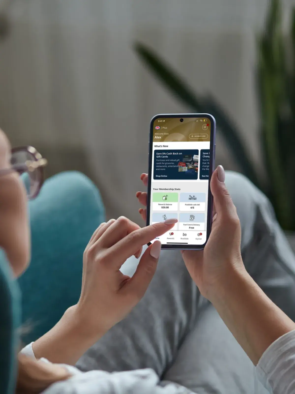 Person relaxing in a chair while using a smartphone displaying a membership rewards and benefits app, with a blurred indoor background and houseplant visible behind.