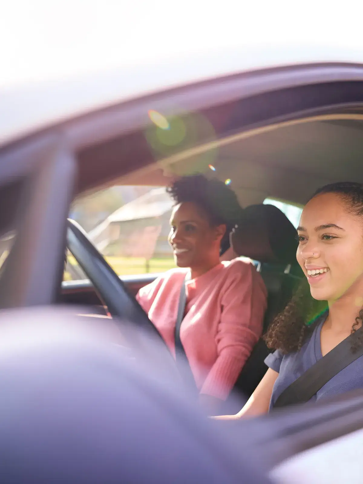 Person driving a car with a passenger seated beside them, both wearing seatbelts as sunlight streams through the windows.