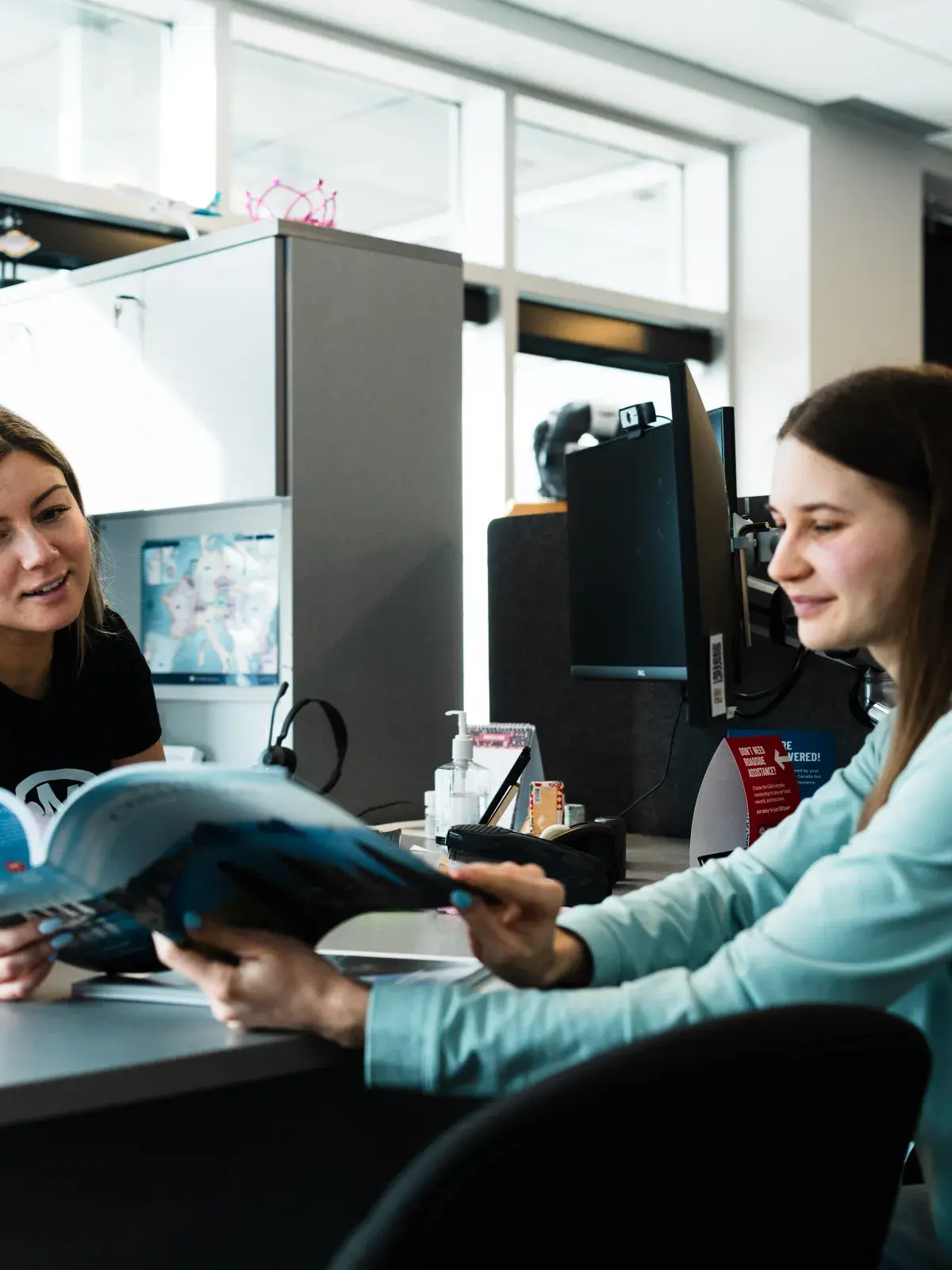 Two people seated at a service desk in an office, reviewing a brochure together beside computer monitors and office equipment.