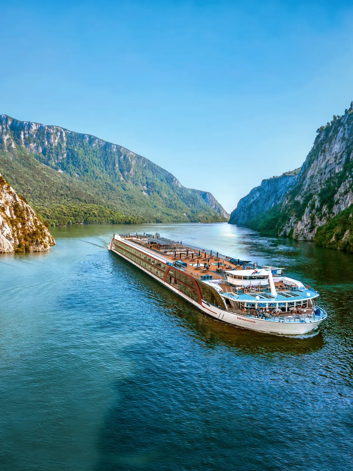AmaWaterways river cruise ship sailing through a scenic river valley surrounded by steep rocky cliffs and green forest under a clear blue sky.