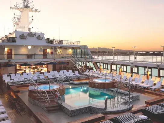 A cruise ship’s open-air deck at sunset, featuring multiple small pools and hot tubs in the center, surrounded by striped lounge chairs. The ship’s mast and upper decks rise in the background, with a distant shoreline silhouetted against a warm orange sky.