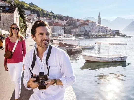 A man with a camera and a woman walk along a scenic European waterfront with boats, stone buildings, and mountains.