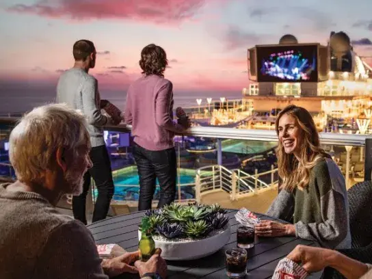 People on a cruise ship deck at sunset, enjoying drinks, playing cards, and watching entertainment on a large outdoor screen.