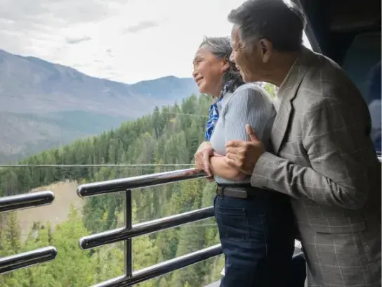Two people stand on a balcony or observation deck, overlooking a scenic view of mountains and forests. The individuals are facing away from the camera, with one person holding the other. The background features lush green trees and distant mountains under a cloudy sky.