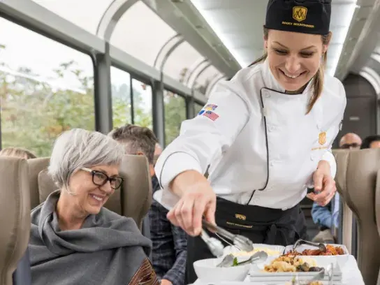 "A chef serves food to passengers on a Rocky Mountaineer train. The chef, wearing a white uniform and black hat, places food onto a plate from dishes on a trolley. Passengers are seated in comfortable chairs, with large windows allowing natural light to enter.