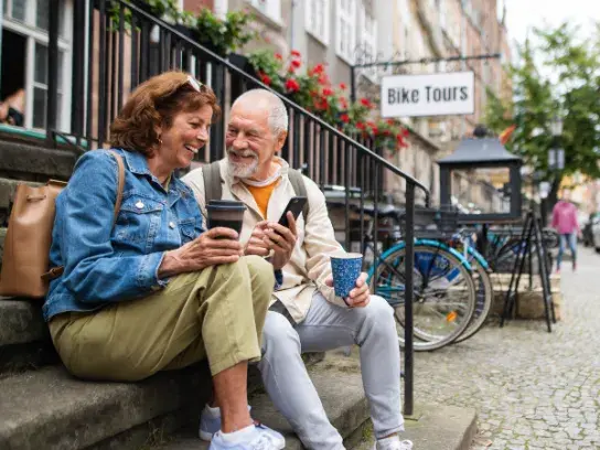 Two people sitting on outdoor steps in an urban area, holding cups and looking at a smartphone. A "Bike Tours" sign is visible in the background, along with bicycles, buildings, and cobblestone pavement.