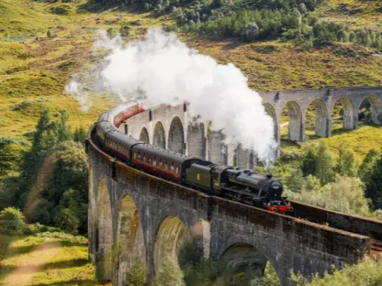A steam train crossing a large, curved stone viaduct with multiple arches, releasing white smoke. The viaduct is surrounded by lush green hills and trees.