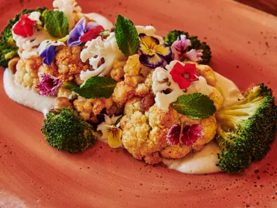 A plated dish featuring roasted cauliflower, broccoli, and edible flowers on a bed of creamy white sauce, garnished with fresh mint leaves. Served in an oval ceramic dish with a rustic, earthy tone.