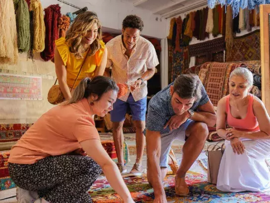 Five people examine a patterned rug in a room with colorful hanging textiles.