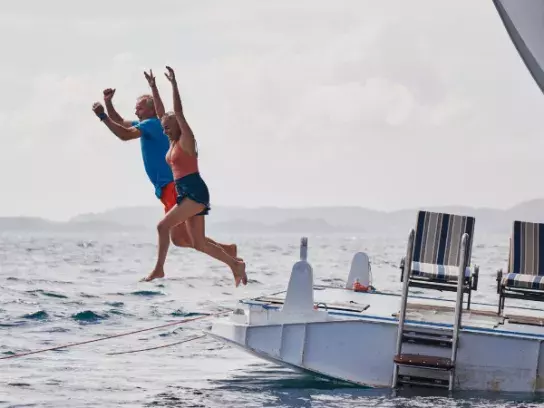 Two people jump off a boat into the water, with striped chairs on the boat's platform and islands in the background.