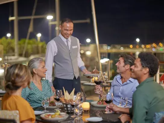 "Five people dining outdoors by a marina at night, with a waiter serving wine."