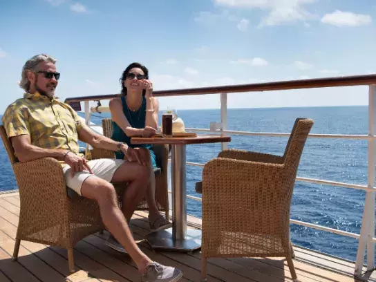 Two people sitting on a boat deck at a small round table, enjoying drinks. The deck has wooden planks and wicker chairs, with the ocean and a partly cloudy blue sky in the background.