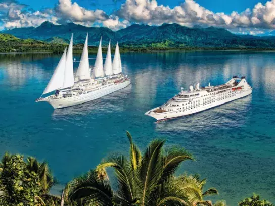 Two large cruise ships sailing on calm blue water—one with white sails and the other a modern liner without sails. Lush green palm trees and tropical vegetation are in the foreground, with green mountains and a partly cloudy sky in the background.