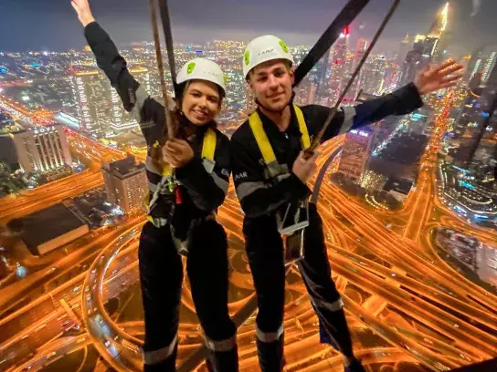 Two individuals wearing safety harnesses and helmets are suspended above a brightly lit cityscape at night. One of them is Travel Advisor Cassidy Royea, enjoying an adventurous activity, possibly zip-lining or bungee jumping.