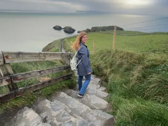 Travel Advisor Shayna walks down a stone staircase toward a coastal path in Ireland, with grassy fields and ocean views under an overcast sky