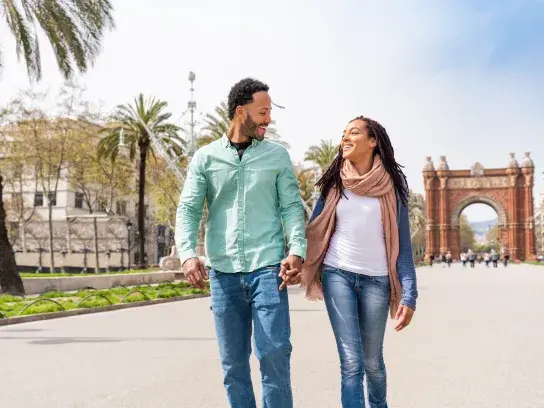 Two people walk hand in hand down a wide street lined with palm trees, with a large red-brick archway in the background. The scene is sunny with clear skies, and the individuals are dressed casually, appearing to be engaged in conversation.