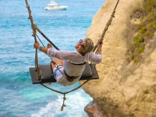 Person wearing sunglasses and a white shirt smiling at the camera while sitting on a swing suspended over a cliff, with the ocean and a distant boat in the background.