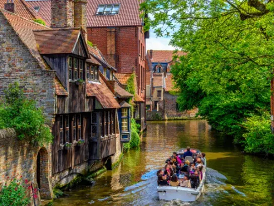 Boat with several passengers cruising along a scenic canal lined with historic brick and timber buildings, surrounded by lush trees and greenery.