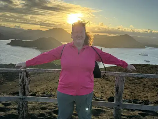 Trish standing with arms outstretched against a wooden railing, overlooking a coastal landscape with island hills and ocean below at sunset.