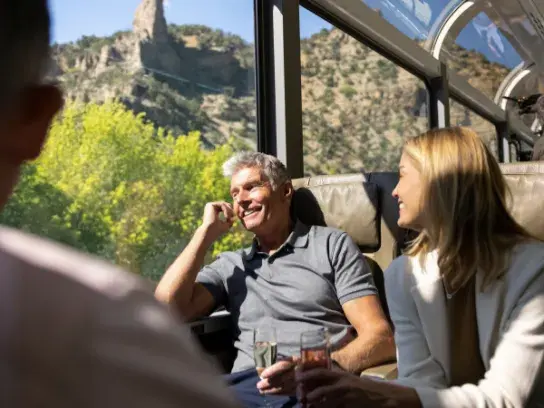 Passengers relax in a scenic train car with large windows, enjoying drinks while mountain views pass by outside.