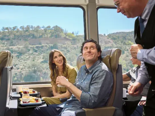 Guests sit in a spacious train cabin with panoramic windows, being served food and drinks as they take in the surrounding landscape.