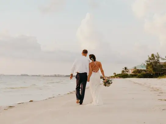 Two people walking away along a sandy beach shoreline, dressed in formal wedding attire, one holding a bouquet. The ocean is on the left, with light waves, and houses and trees line the right side. Soft evening light and clouds fill the sky.