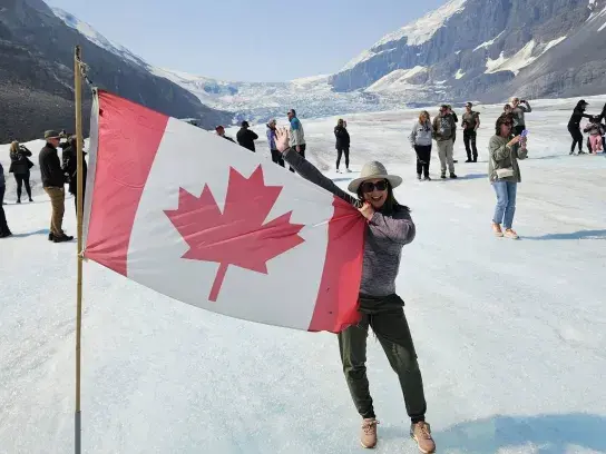 A person stands on a glacier holding a large Canadian flag while surrounded by other visitors. Snow-capped mountains and an expansive ice field stretch into the distance under a clear blue sky. The flag fills much of the foreground, with the glacier and mountain landscape providing a dramatic backdrop.