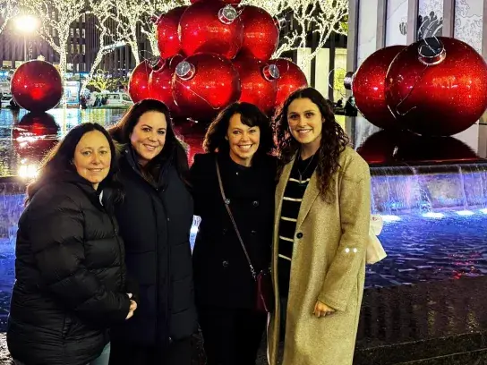 Four travellers posing in front of oversized red holiday ornaments and illuminated city lights at night.