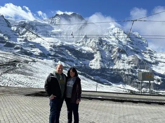 Two travellers standing at a mountain viewpoint with snow-covered peaks and glaciers under a bright blue sky.