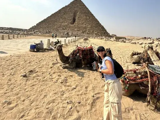 Kailyn standing beside resting camels in front of the Great Pyramid of Giza on a sunny day, with desert sand and historic stone structures in the background.