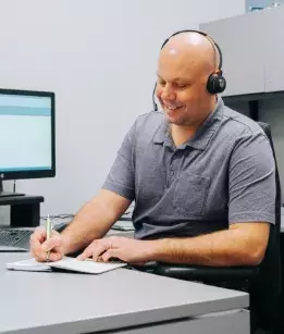 Man wearing a grey polo shirt sitting at a desk with a headset on writing on a piece of paper with a computer in the background