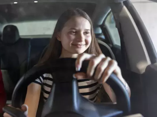 Young woman smiling and confidently driving a car, viewed from the interior with a focus on her hands on the steering wheel.