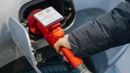 Person in a dark jacket refueling a car with a red fuel pump, featuring a Shell GO+ sign