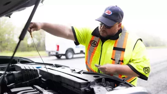 Roadside assistance worker in a bright saftey vest checking under the hood of a car on a highway shoulder with a white truck in the background