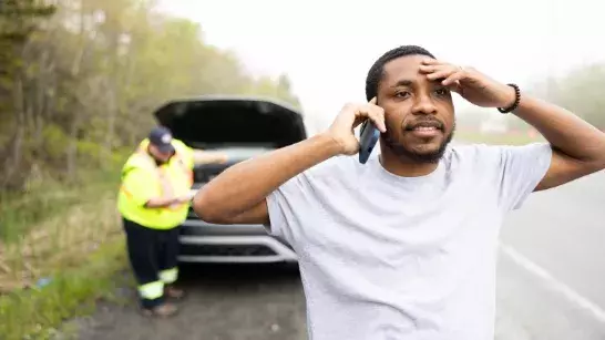 Man wearing white tshirt on the shoulder of a highway looking a bit distressed while on the phone with his hand on his forehead. Roadside assistance worker wearing neon safety gear under the roof of car in the background