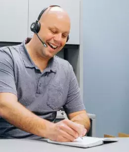 bald gentleman wearing a blue polo tshirt with a headset smiling while writing on a notepad