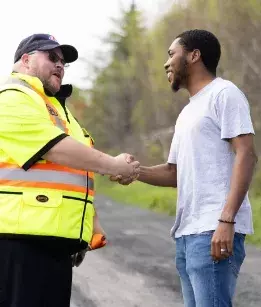 Male roadside assistance worker in neon short sleeve jacket shaking hands with a male wearing a grey tshirt and jeans smiling