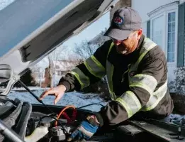 Roadside assistance worker wearing a ball cap and safety gear working under the hood of a vehicle 