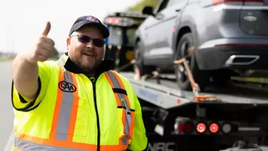 close up shot of a roadside assistance worker wearing neon reflective gear, sunglasses and a CAA hat giving the thumbs up with a car secured to a tow truck behind him
