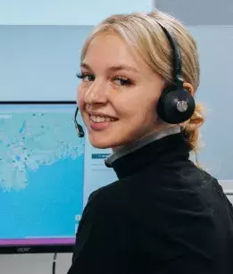 Woman wearing a headset and a black turtleneck turning her head back over her shoulder smiling with a computer screen in the background