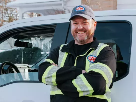 CAA Roadside Assistance worker in reflective gear smiling wiht crossed arms leaning up against a truck