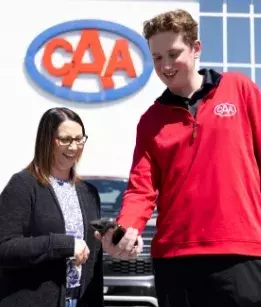 tall male CAA worker in a red shirt showing a shorter female wearing a black sweater and glasses something on his phone with the CAA logo on the side of a building in the background