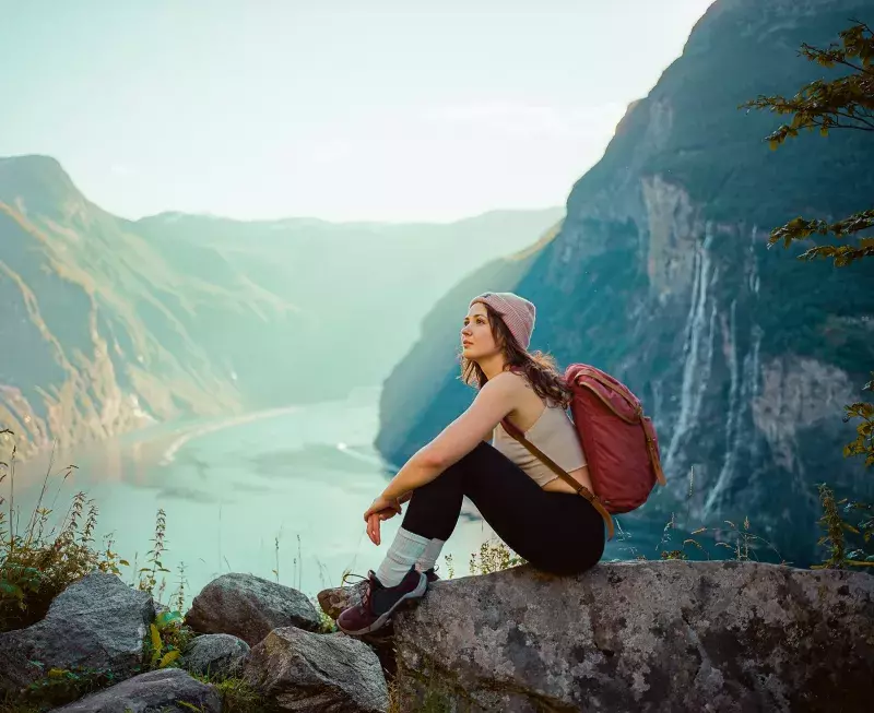 A person wearing a beanie, tank top, leggings, and hiking shoes sits on a rock with a backpack. They are in a mountainous area with a river and waterfall in the background, looking contemplatively into the distance.