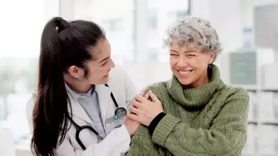 Female doctor wearing a white coat with long hair in a ponytail smiling with her hand on the shoulder of an older lady wearing a green sweater 