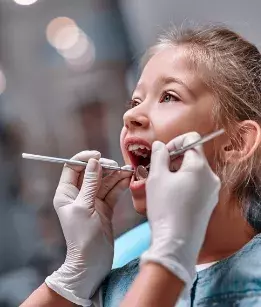 Two hands wearing medical gloves holding up dental tools to a childs open mouth