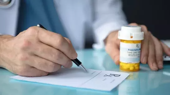 Close up shot of a hand holding a pen writing on a piece of paper that says RX and other hand is holding a medication bottle