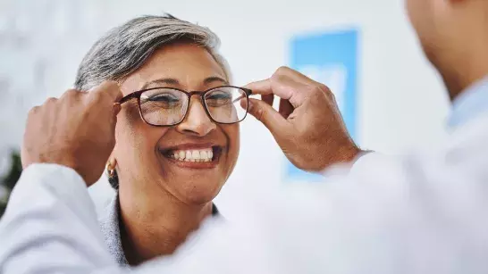 middle-aged Lady smiling while an eye doctor puts a pair of black-framed glasses on her 