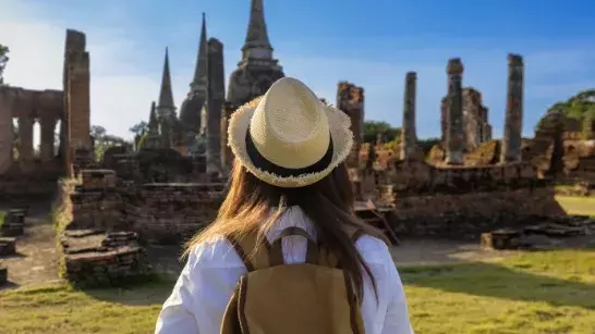 Young woman looking at ruins wearing a white shirt, sun hat and a brown backpack